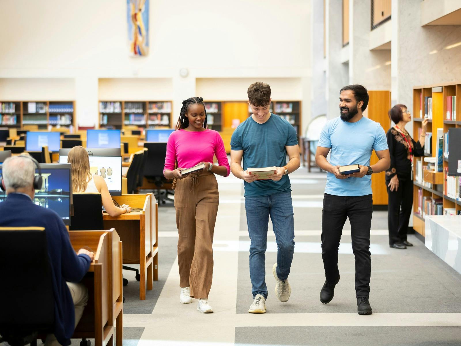 Three young adults walk down a bright, modern library aisle lined with bookshelves.