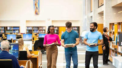 Three young adults walk down a bright, modern library aisle lined with bookshelves.