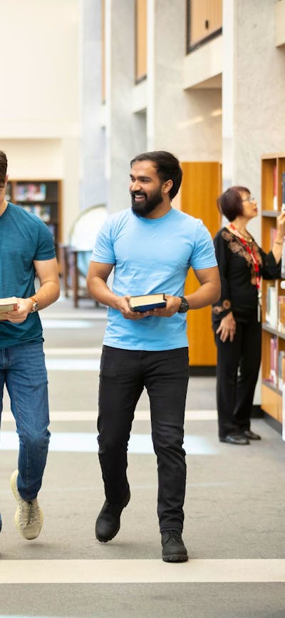 Three young adults walk down a bright, modern library aisle lined with bookshelves.