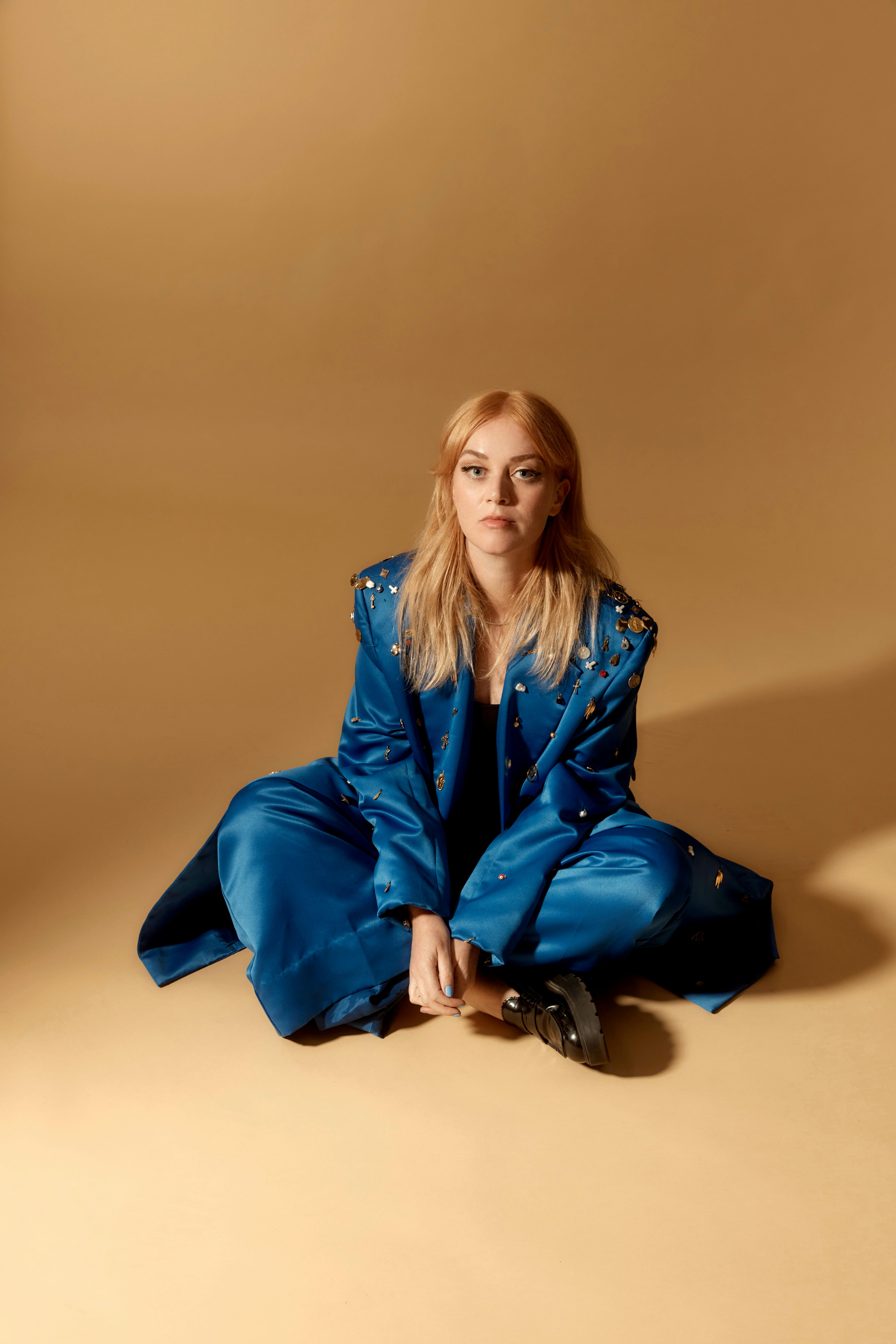 Ella Thompson on the floor in a bright blue embellished suit agains a warm beige studio backdrop