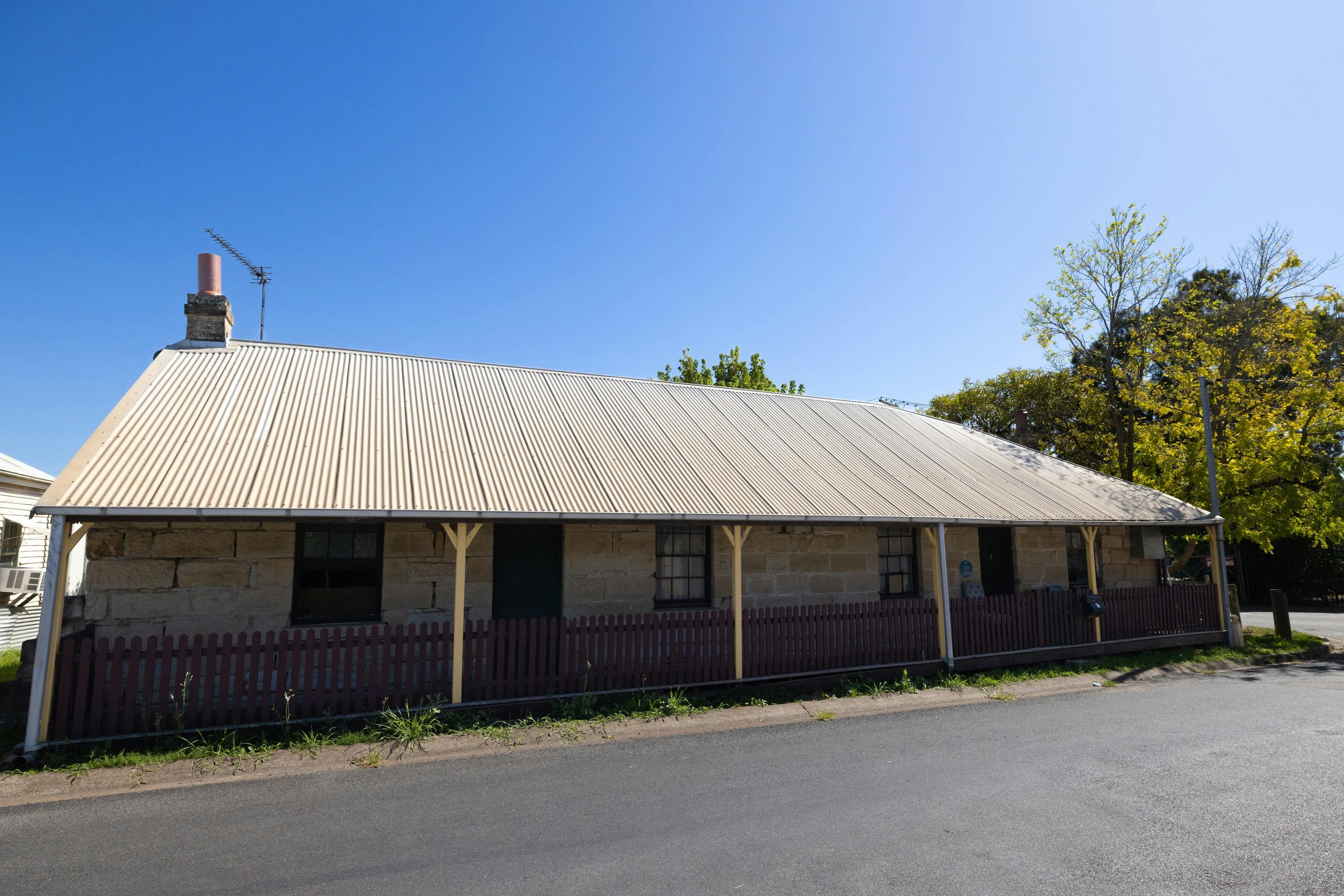 Caroline Chisholm Cottage from the front, sandstone brick, maroon slat fence, corrugated, blue sky