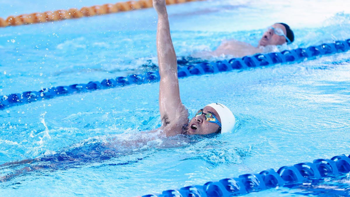 A swimmer executes the backstroke in pool, with another competitor visible in the lane behind.