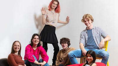A group of six smiling young people aged 12–18 sit on bean-bags and chairs, facing the viewer.