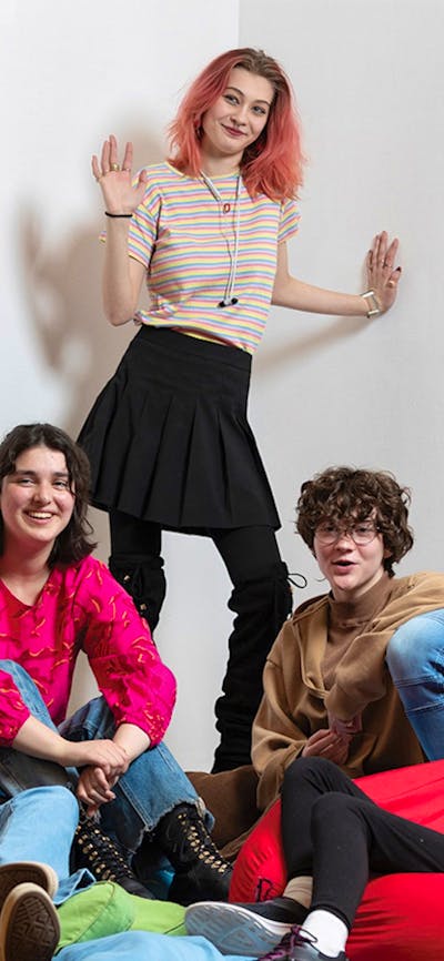 A group of six smiling young people aged 12–18 sit on bean-bags and chairs, facing the viewer.