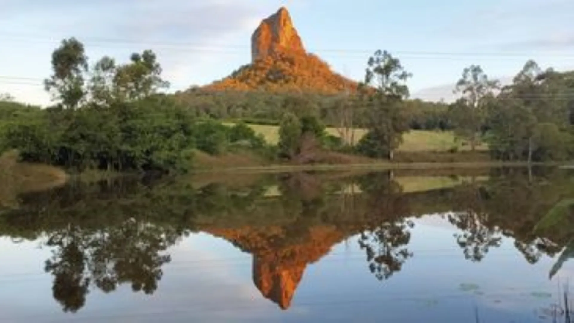 Image shows Mt Coonowrin with late afternoon sun shining, and reflected in a lake in the foreground