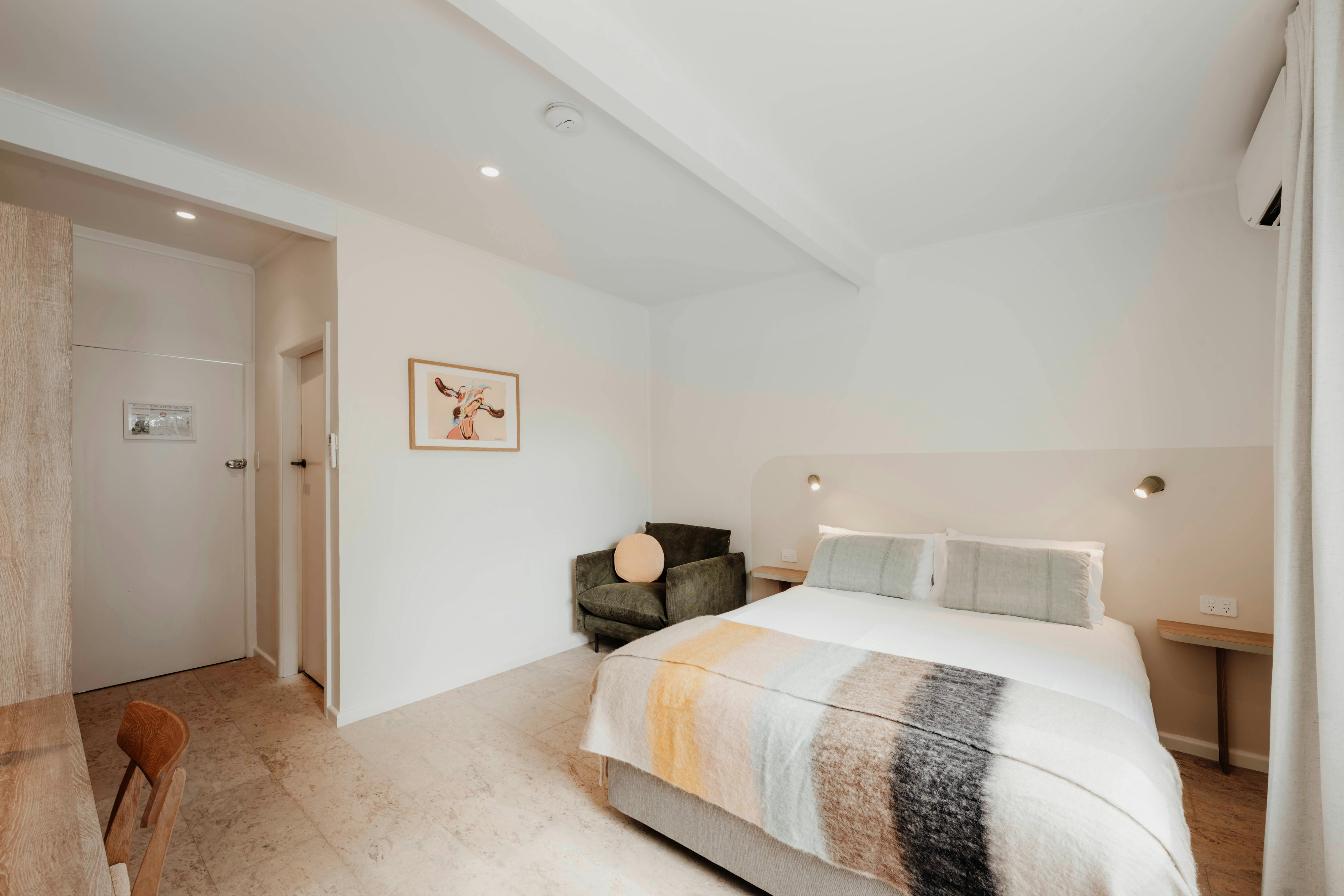 Queen bed with crisp white sheets and stripy blanket. Arm chair and lighting. Art on wall.