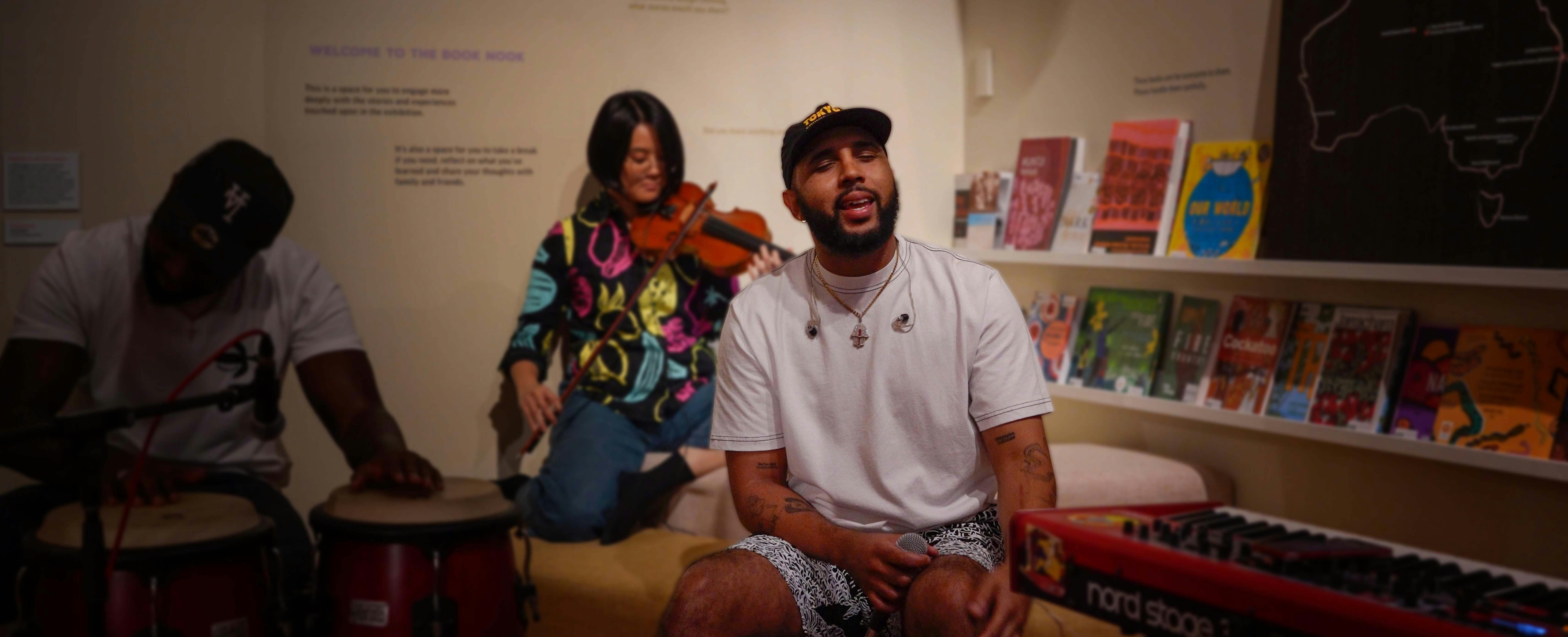 Man singing with his eyes closed in a museum setting, woman playing a violin in the background