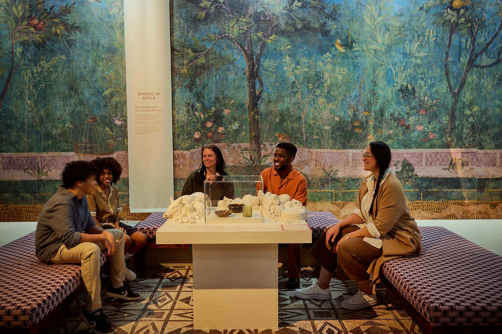 Group of visitors seated around a table exhibit, with a painted Roman garden mural background