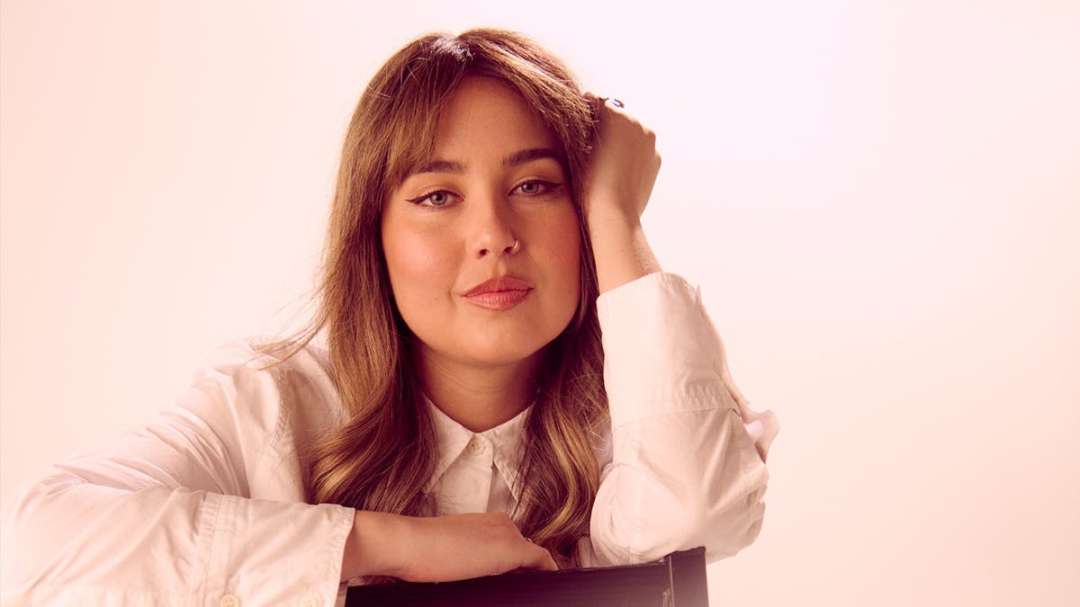 A young woman with long brown hair, sitting back to front on a chair with her left hand rasied