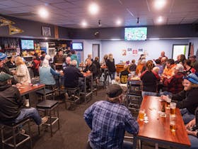 The front bar of a pub, with patrons happily congregating at high tables. A sports match plays on TV