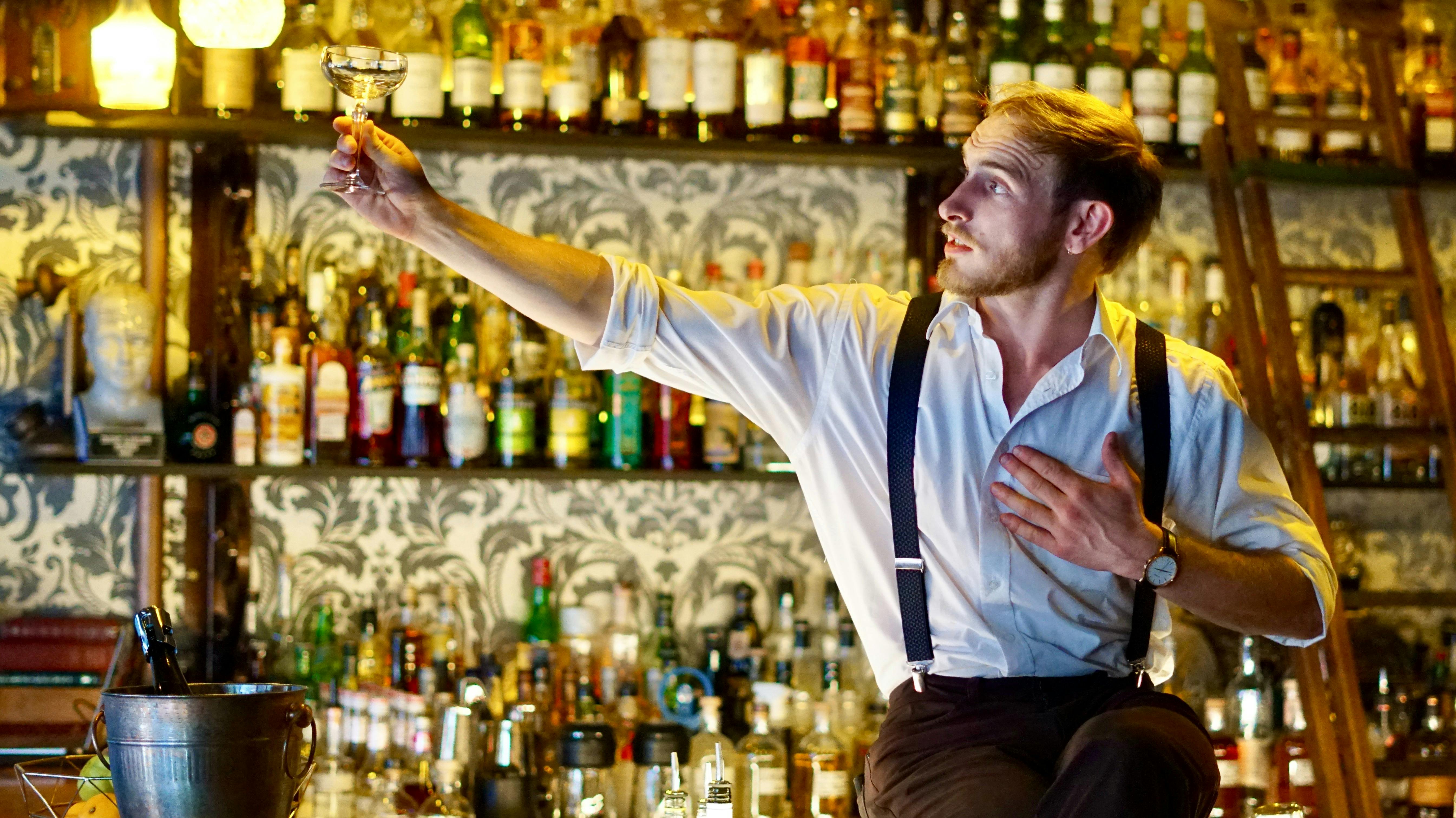 A bartender/performer sits on the bar, holding a glass to the light