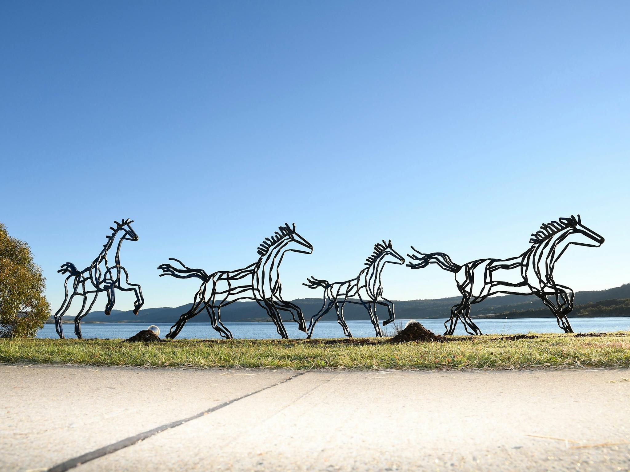 Metal horse sculptures in motion by a lake, set against a clear blue sky.