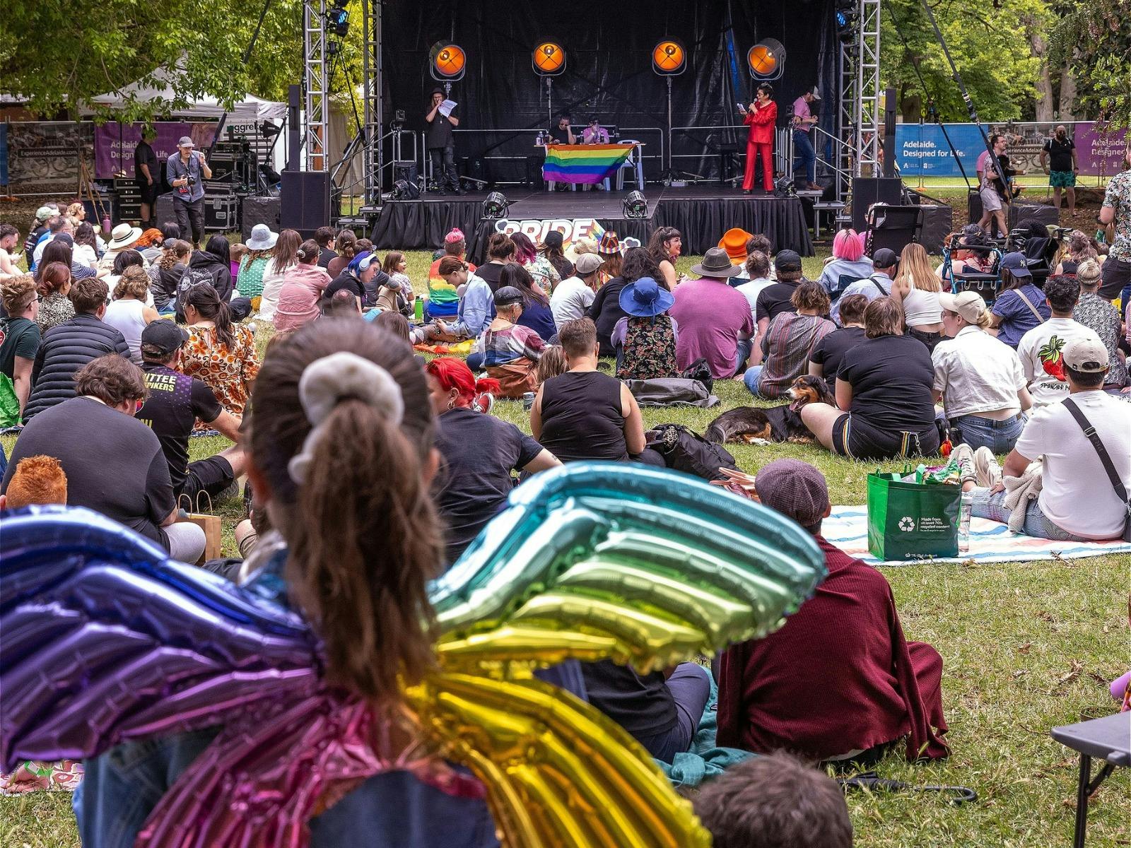 Crowd of people sitting on grass looking towards a stage with pride flags displayed.