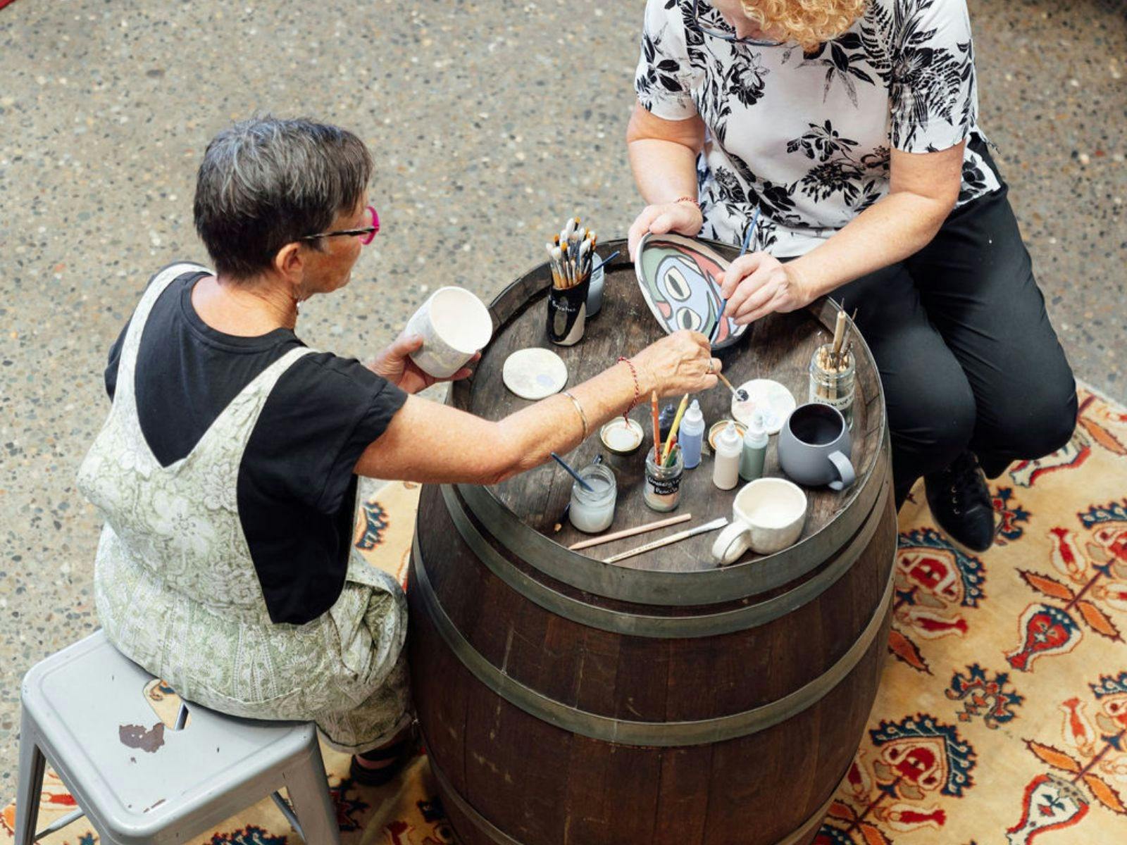 two females sitting at a wine barrel table painting a ceramic piece