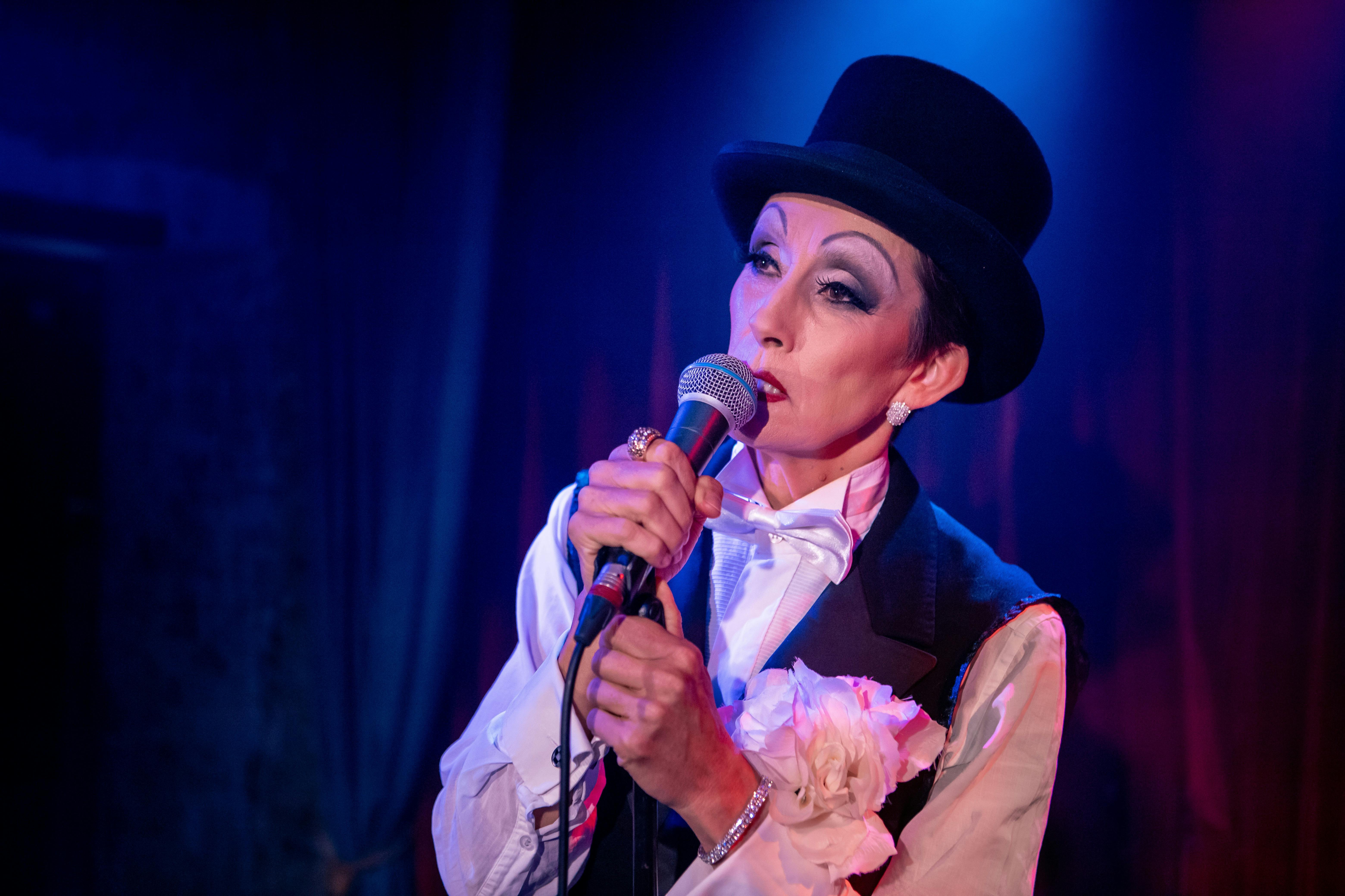 Woman dressed in a tuxedo wearing a top hat, onstage holding mic