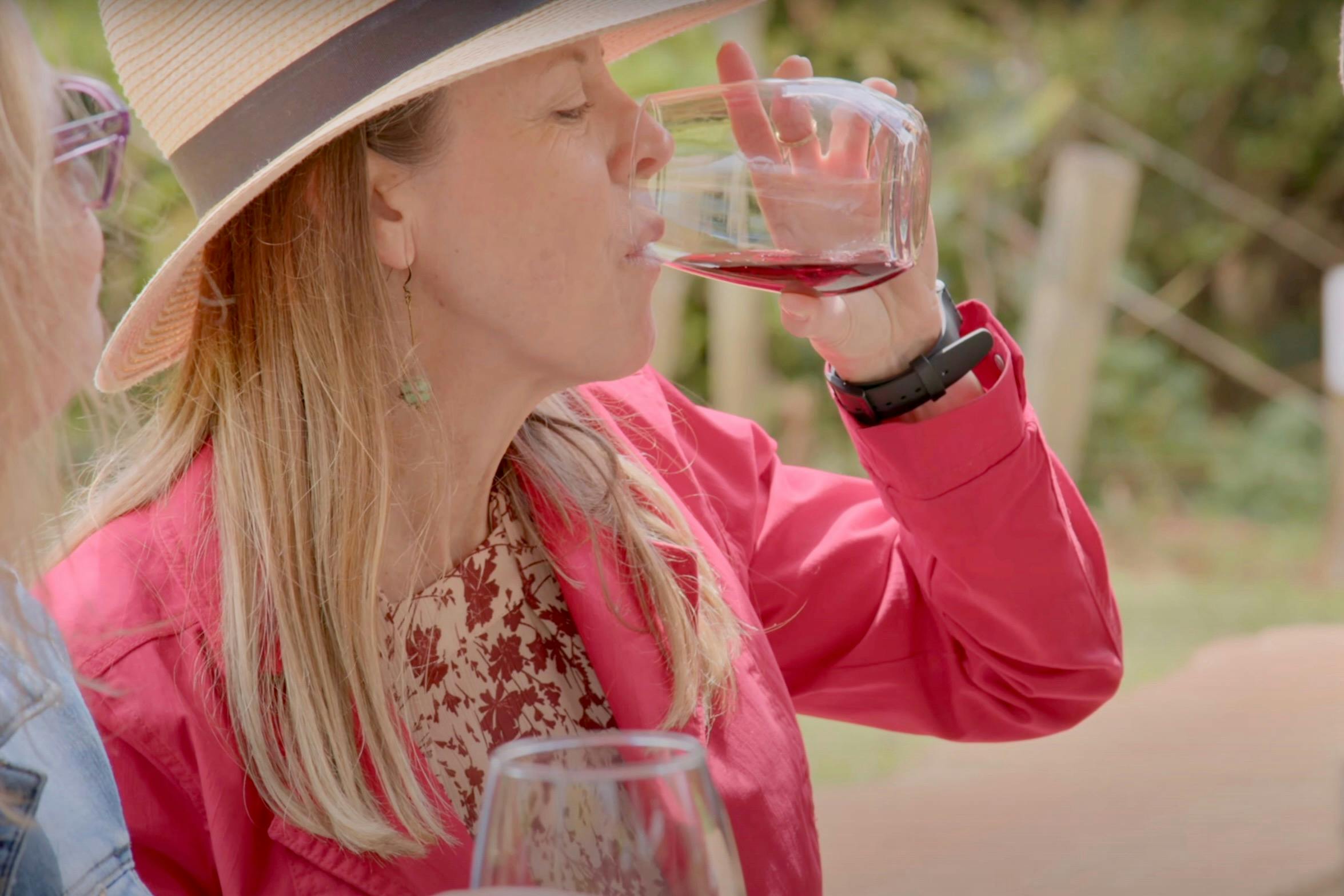 A woman wearing a red jacket and straw hat sipping a glass of red wine