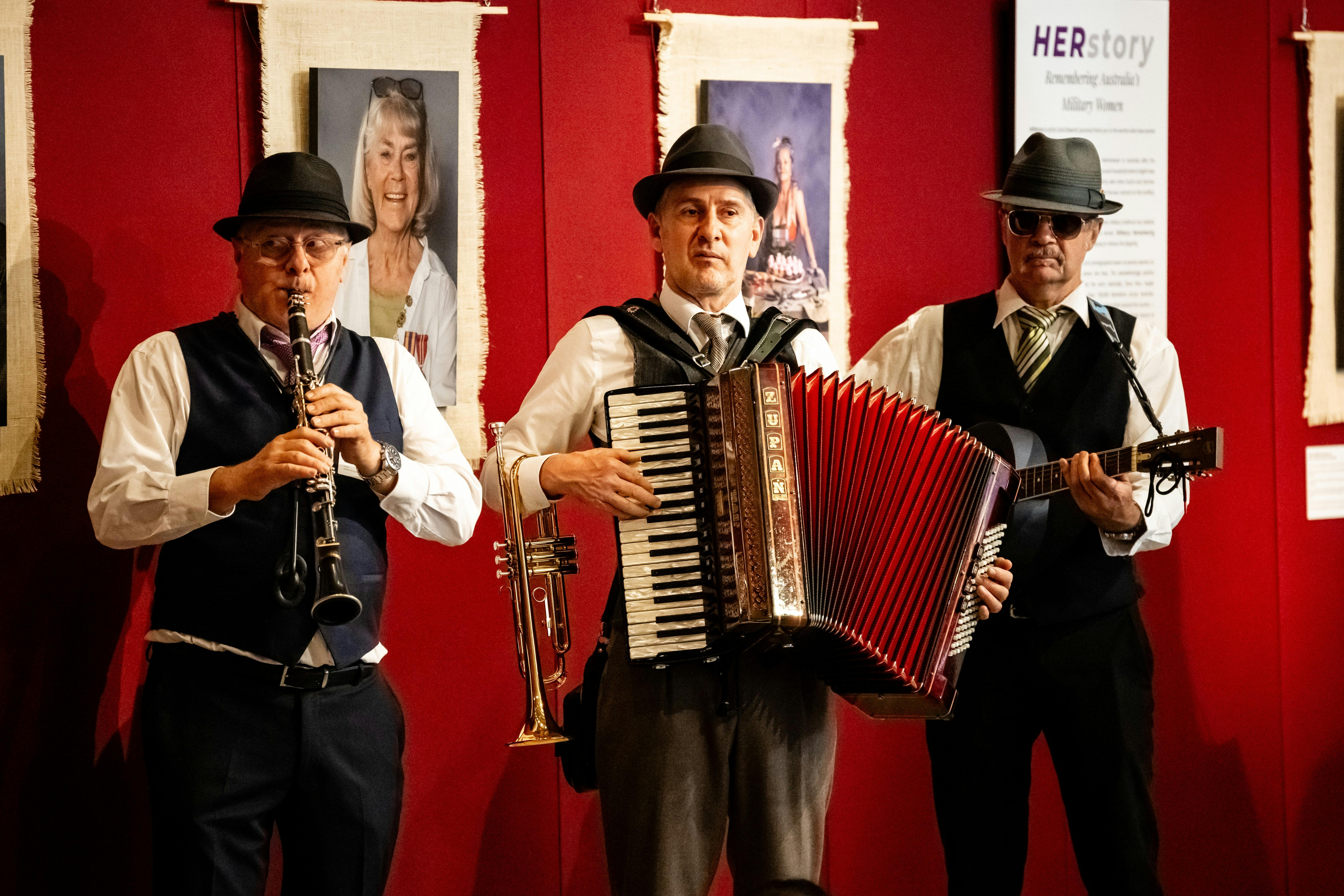 3 musicians wearing shirts, ties, waistcoats and trilby hats play the clarinet, accordian and guitar