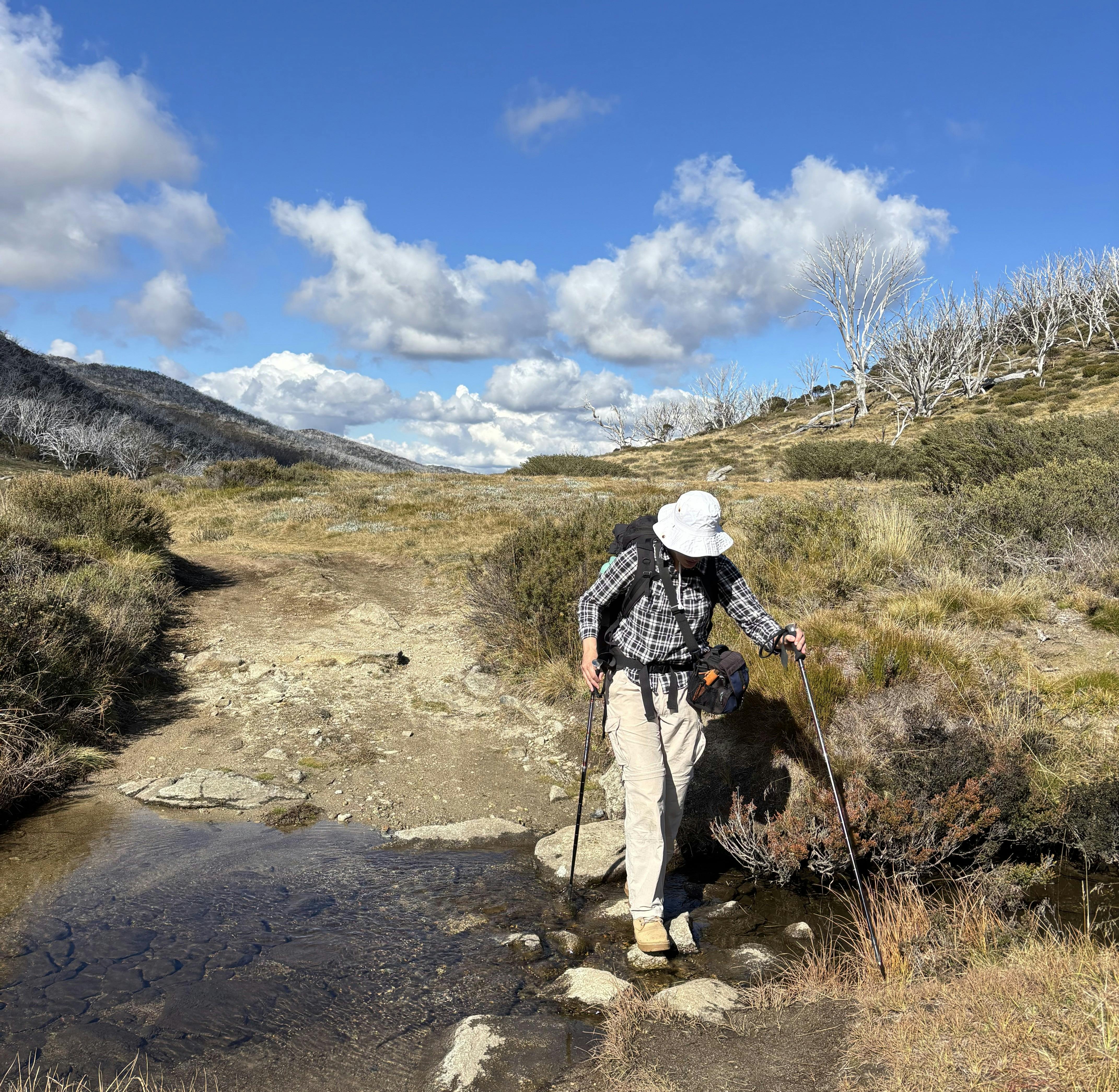 A woman wearing a checkered shirt and white trousers crossing a mountain stream.