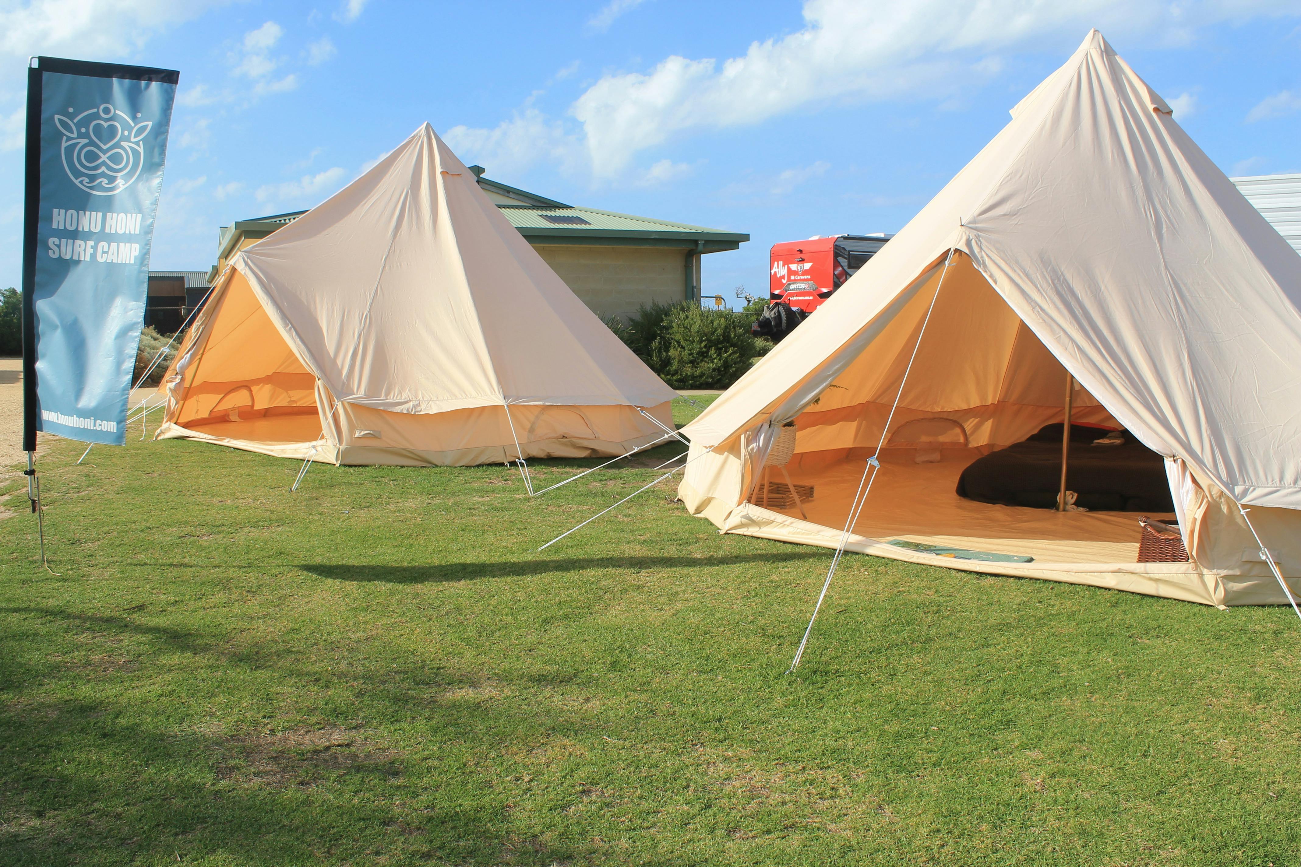 Two bell tents - glamping tents - set up on a grass patch on a sunny day. Honu Honi Surf Camp flag.