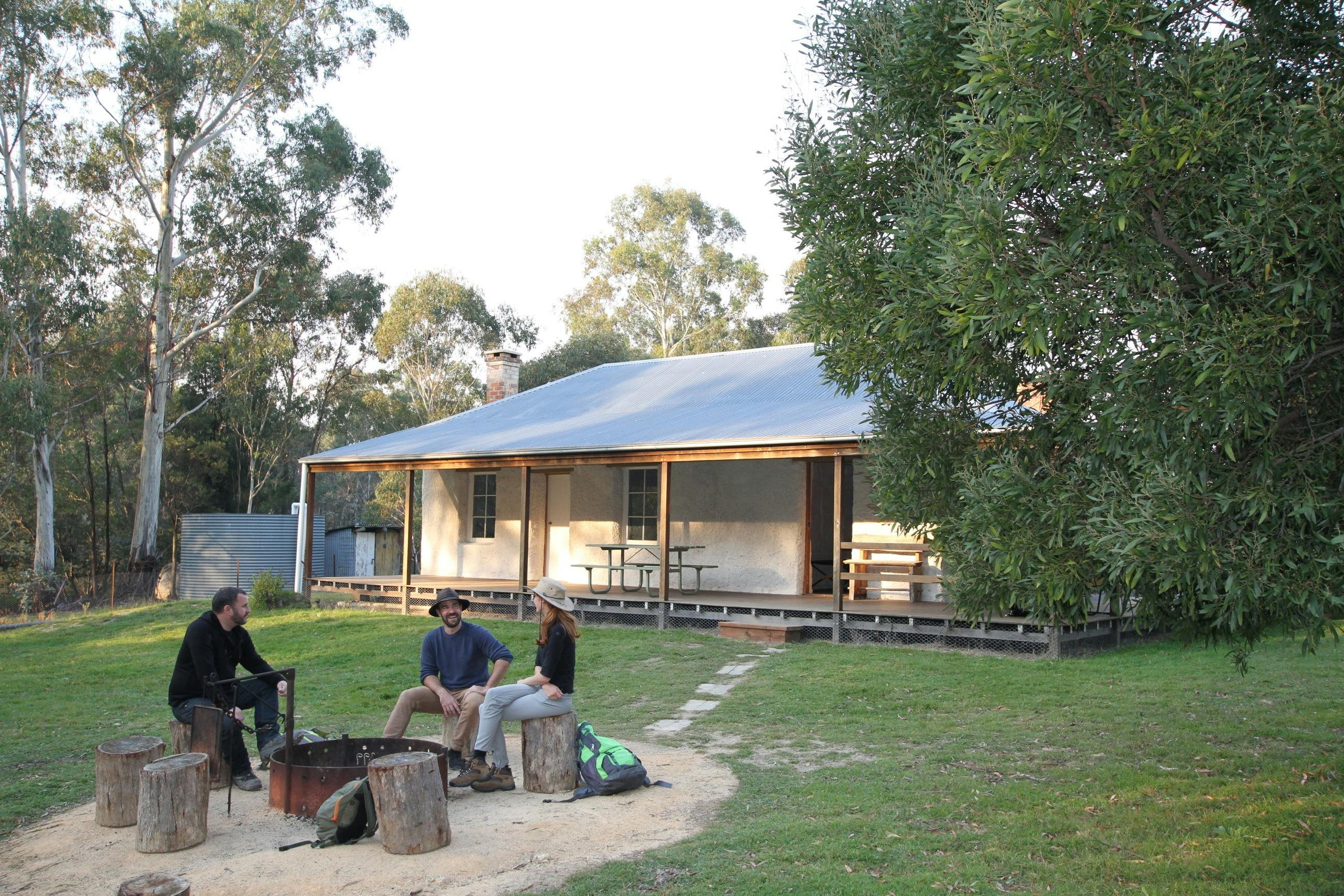 3 people sitting around a firepit among green grass and Heritage property in background