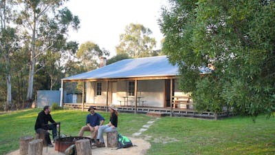 3 people sitting around a firepit among green grass and Heritage property in background