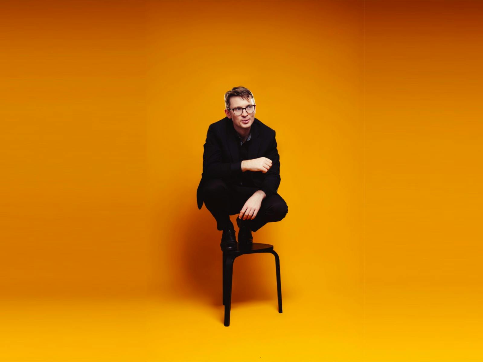 Magician Sam King, dressed in black, crouches on a stool against a bright orange background.