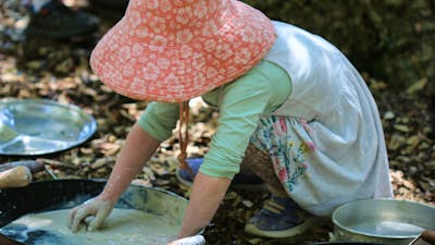 A young child wearing a sunhat, mixing dirt and water in an old frying pan