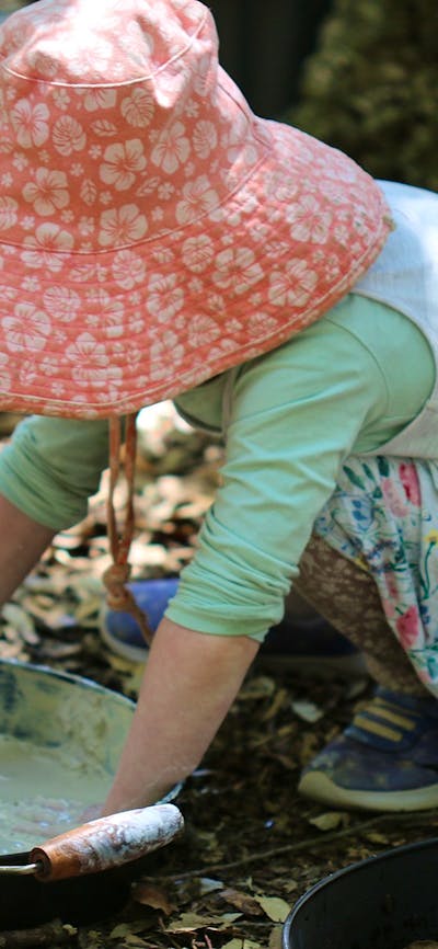 A young child wearing a sunhat, mixing dirt and water in an old frying pan