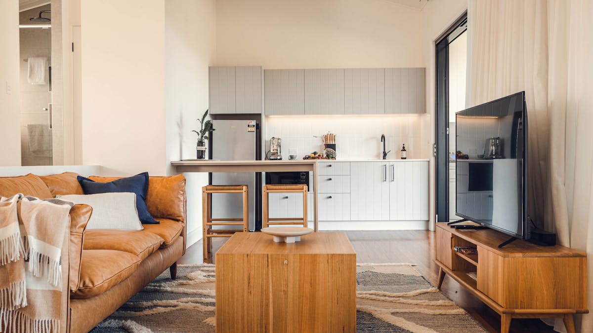 Living area with tan leather lounge, coffee table on rug and large tv with Kitchen in background