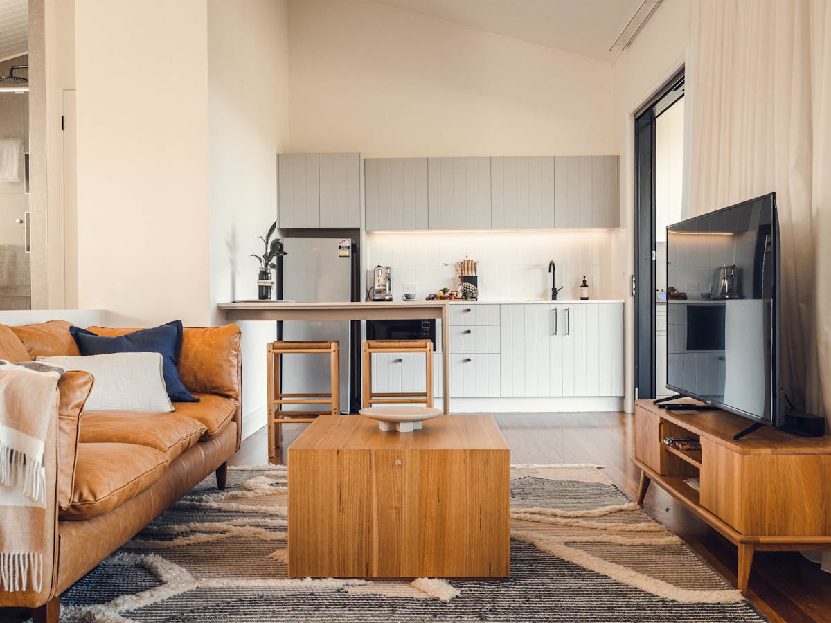 Living area with tan leather lounge, coffee table on rug and large tv with Kitchen in background
