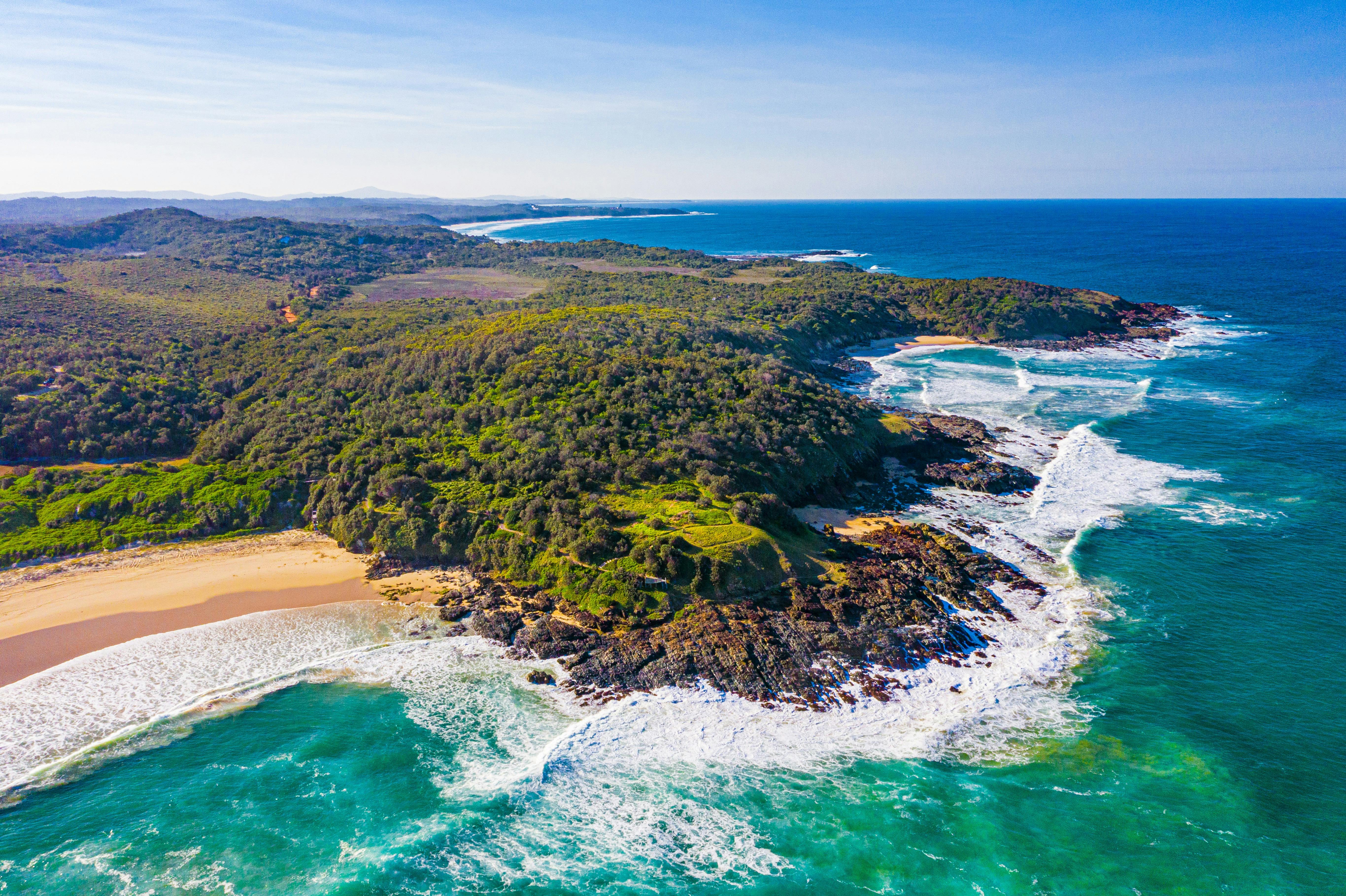 Ariel view of a beach and bush covered headland and blue ocean.
