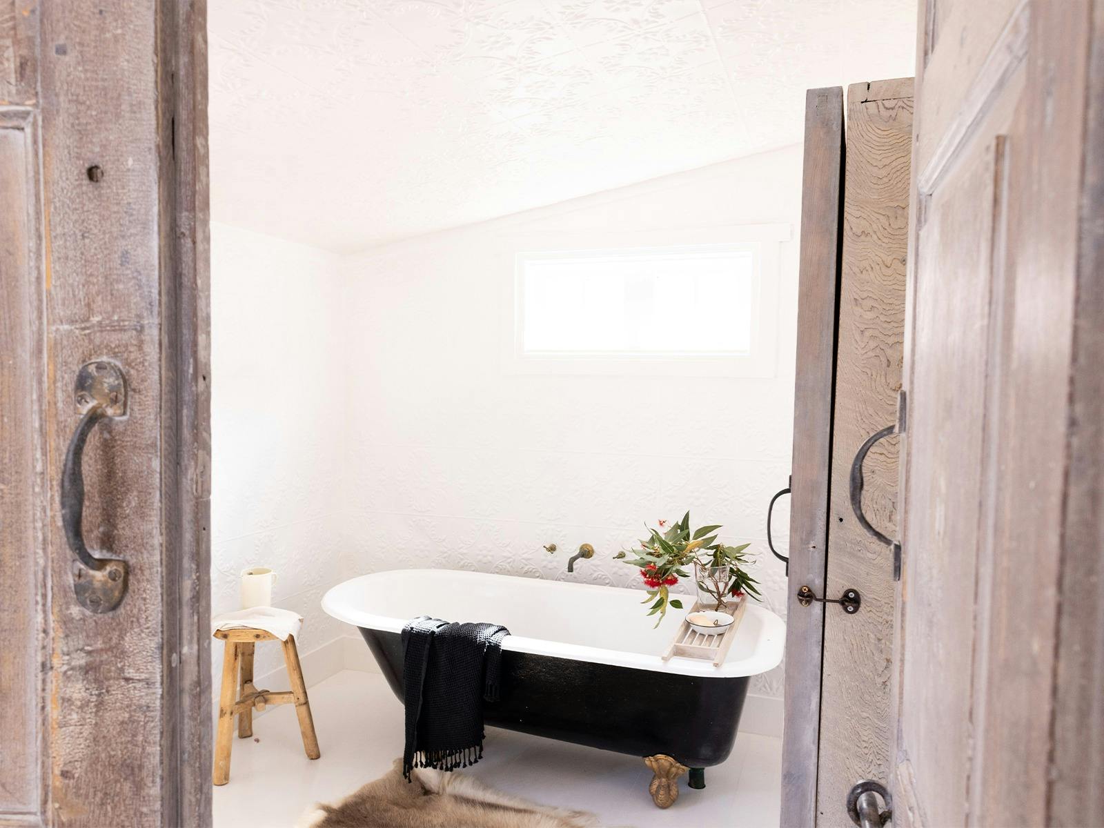 A black claw foot bath tub sits in an all white pressed tin bathroom with wooden details