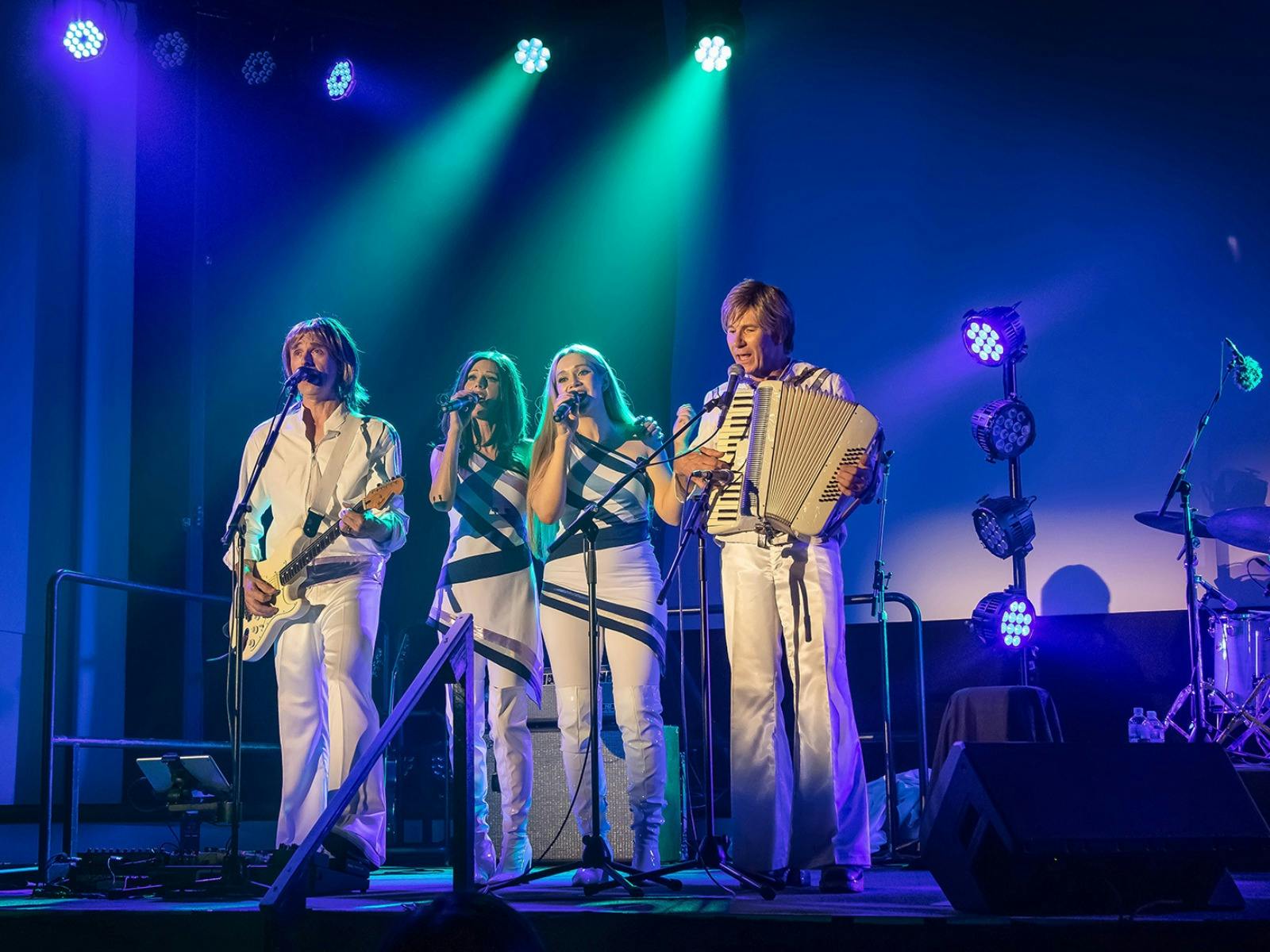 a group of 4 people performing in ABBA style costumes on a stage with lights and microphones