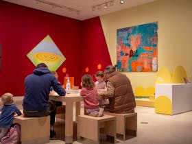 A man, a woman and four young children sit and draw at a table in a colourful gallery