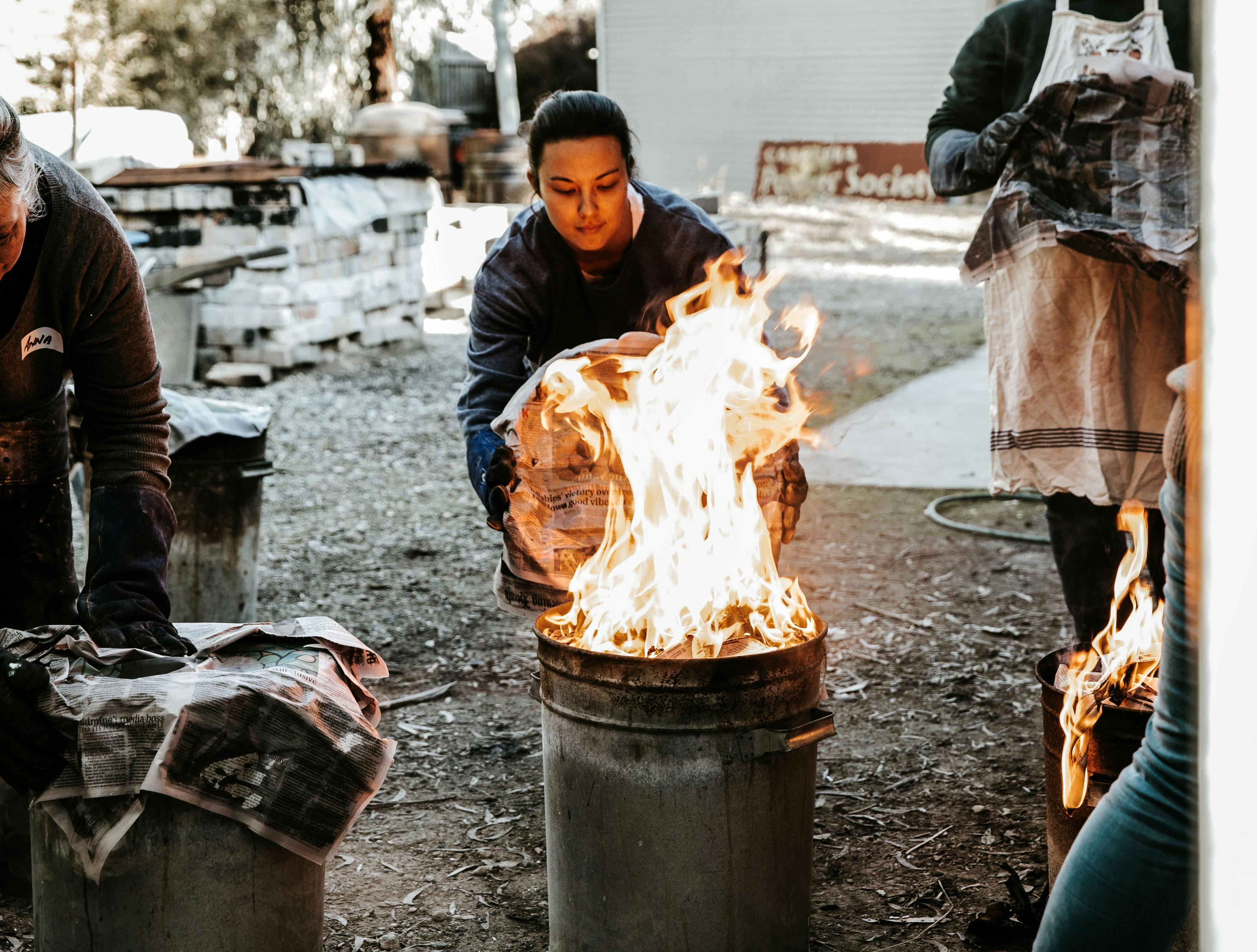 A female student reaches a metal garbage can lid towards a bin full of flames as part of raku firing