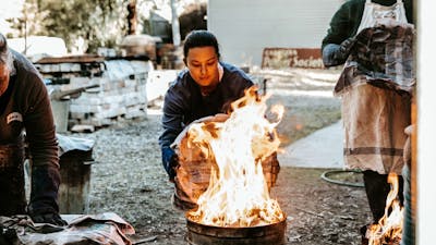 A female student reaches a metal garbage can lid towards a bin full of flames as part of raku firing