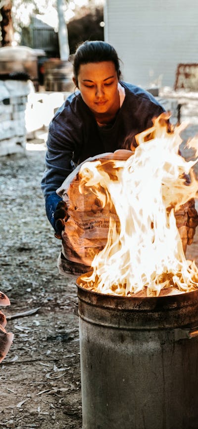 A female student reaches a metal garbage can lid towards a bin full of flames as part of raku firing