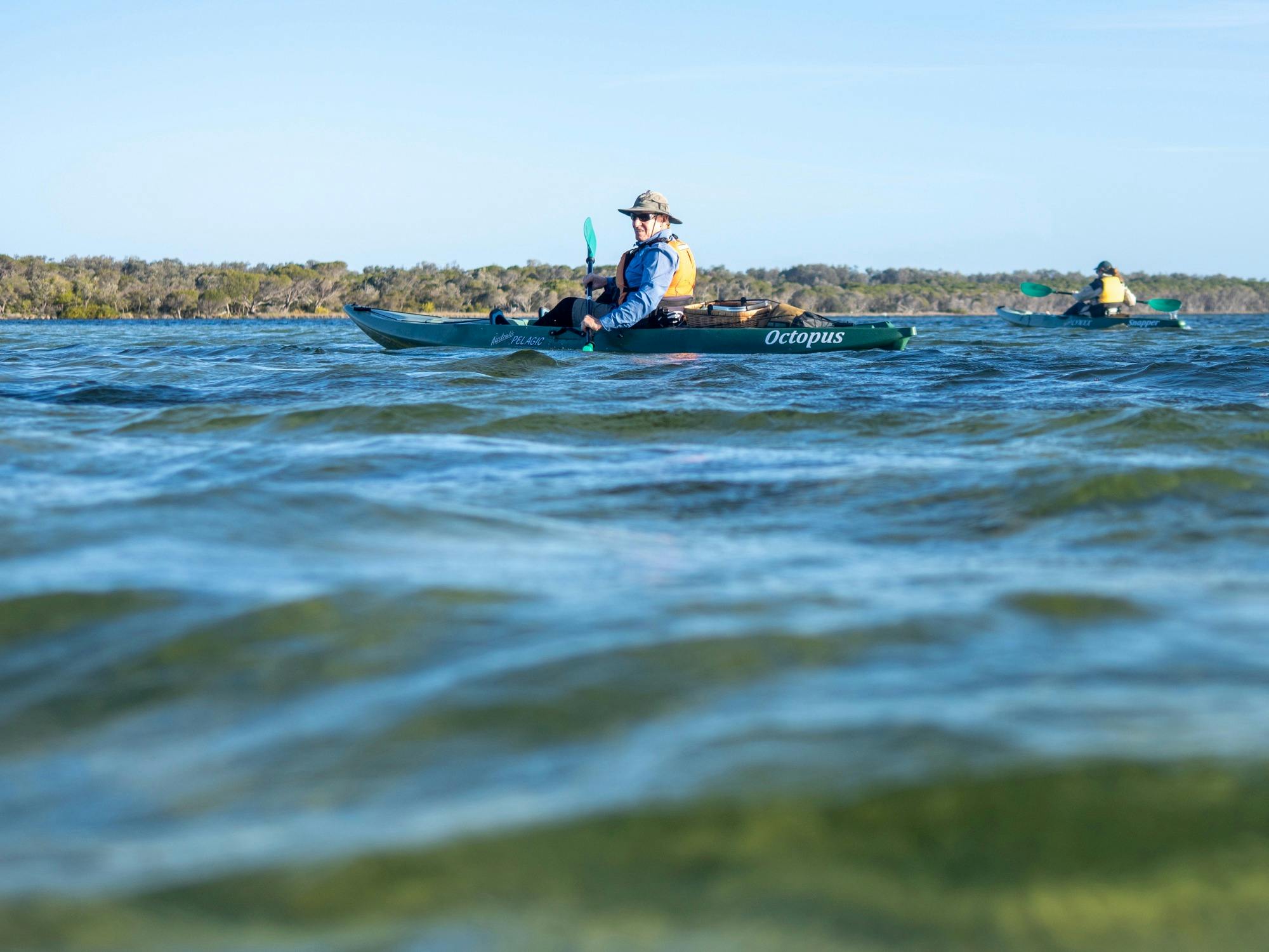 Two kayakers paddling in the distance on a calm blue lake at golden hour.