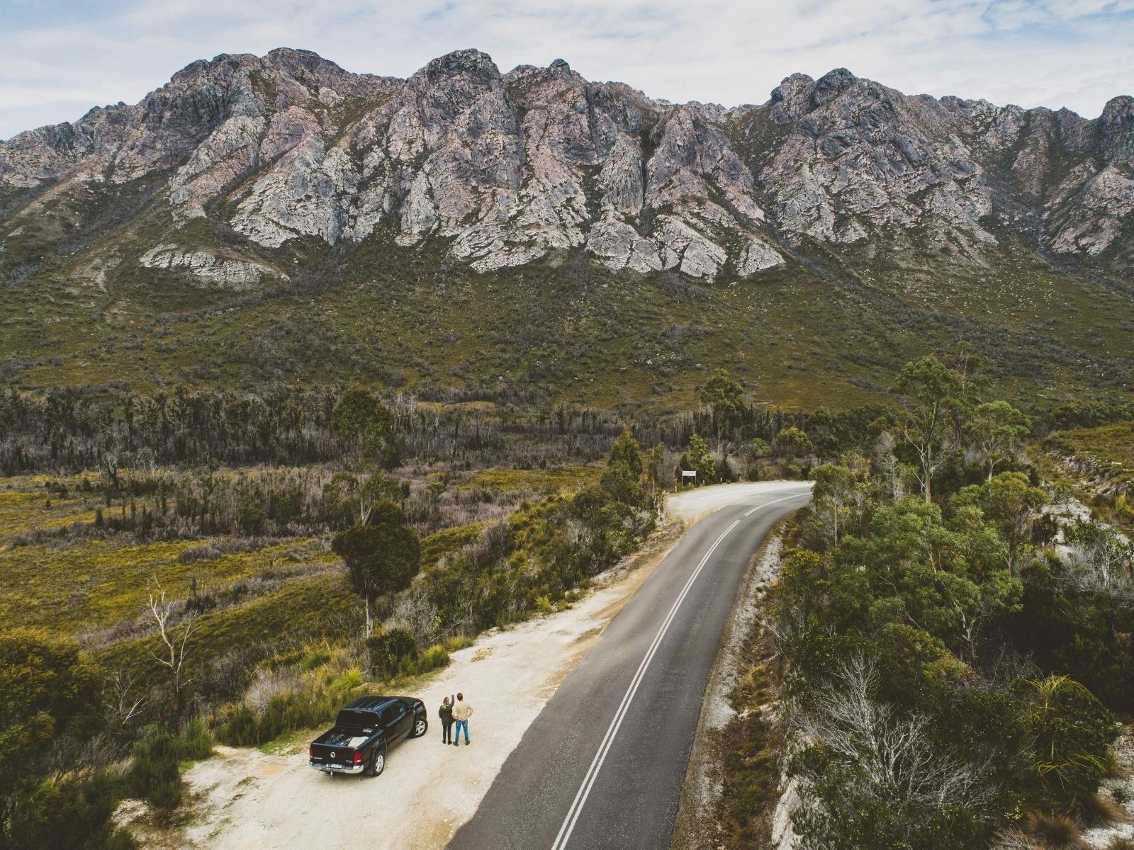 An aerial image of two people standing next to a parked car looking up at a rocky mountain