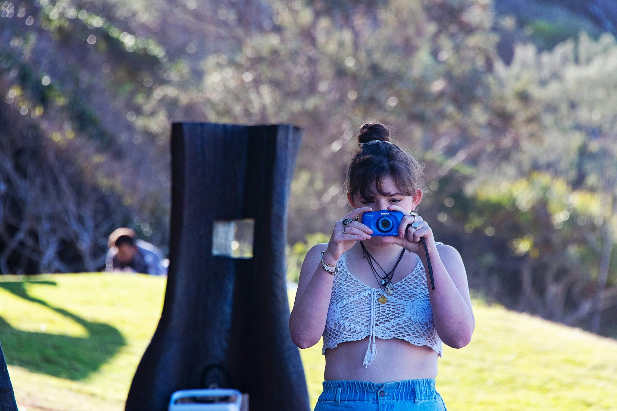 A young woman with a blue point and shoot camera taking a photo with a timber sculpture near her
