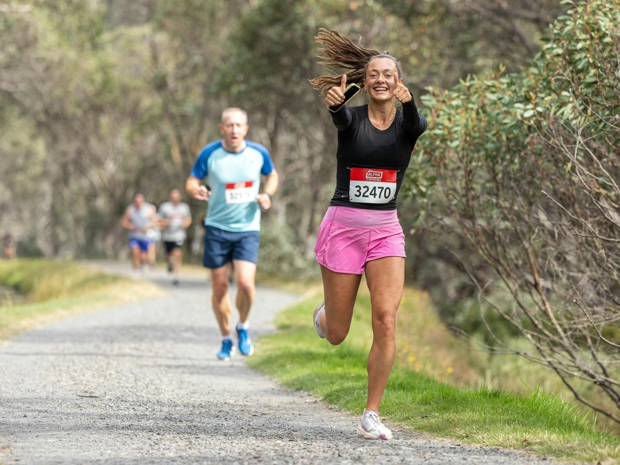 Women giving a thumbs up to the camera on a gravel trail with runners behind her