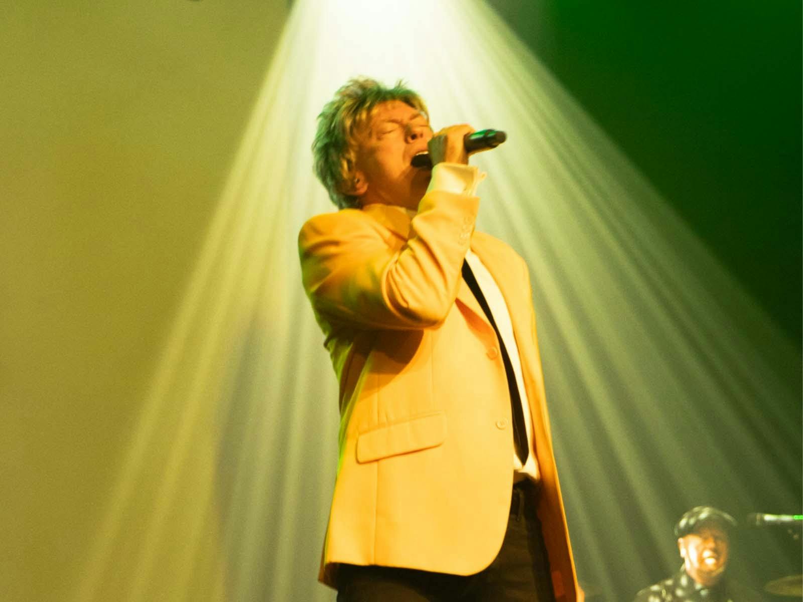 man singing with microphone on stage with yellow jacket