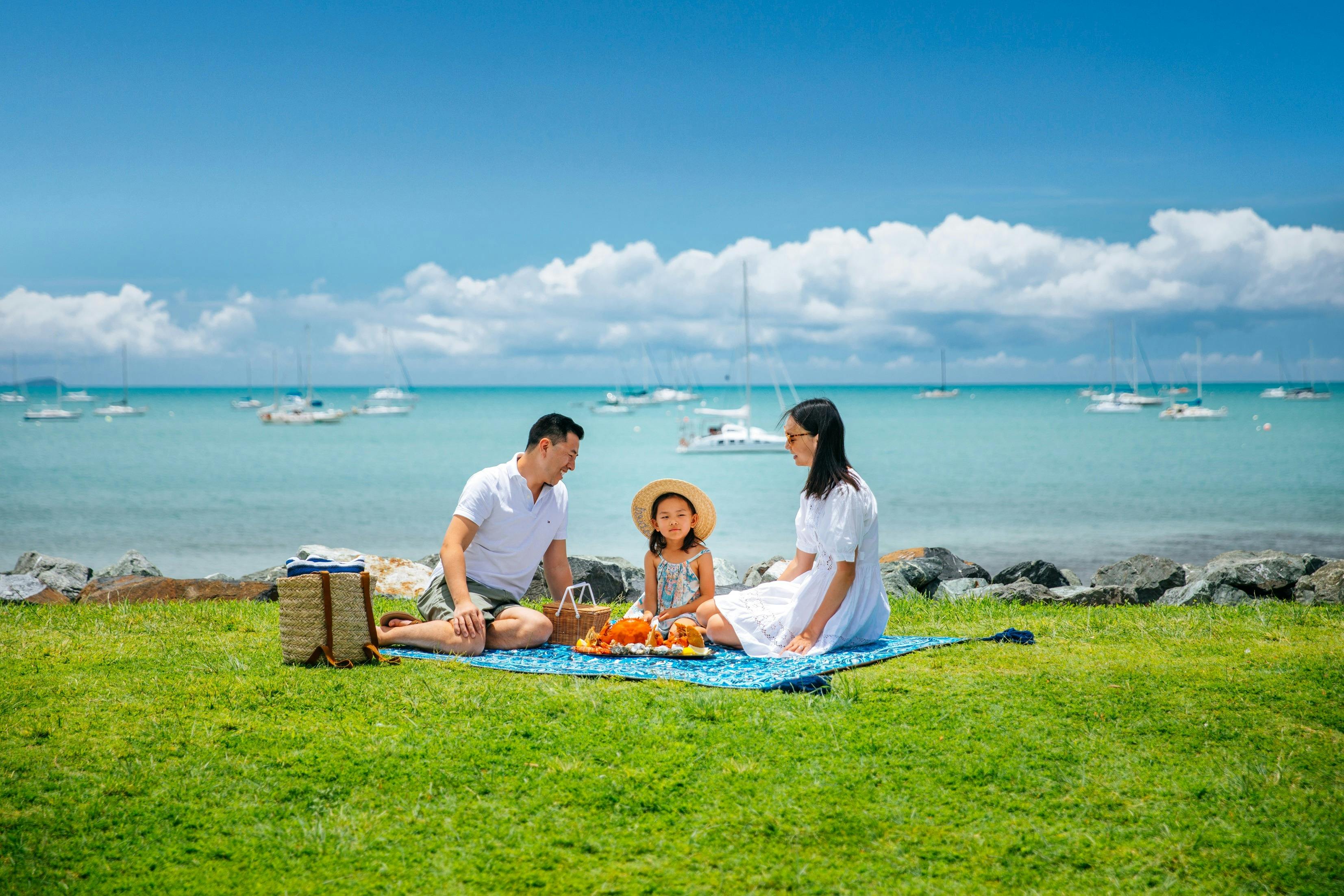 Couple and child sitting having a picnic on grass area with ocean and boats behind them