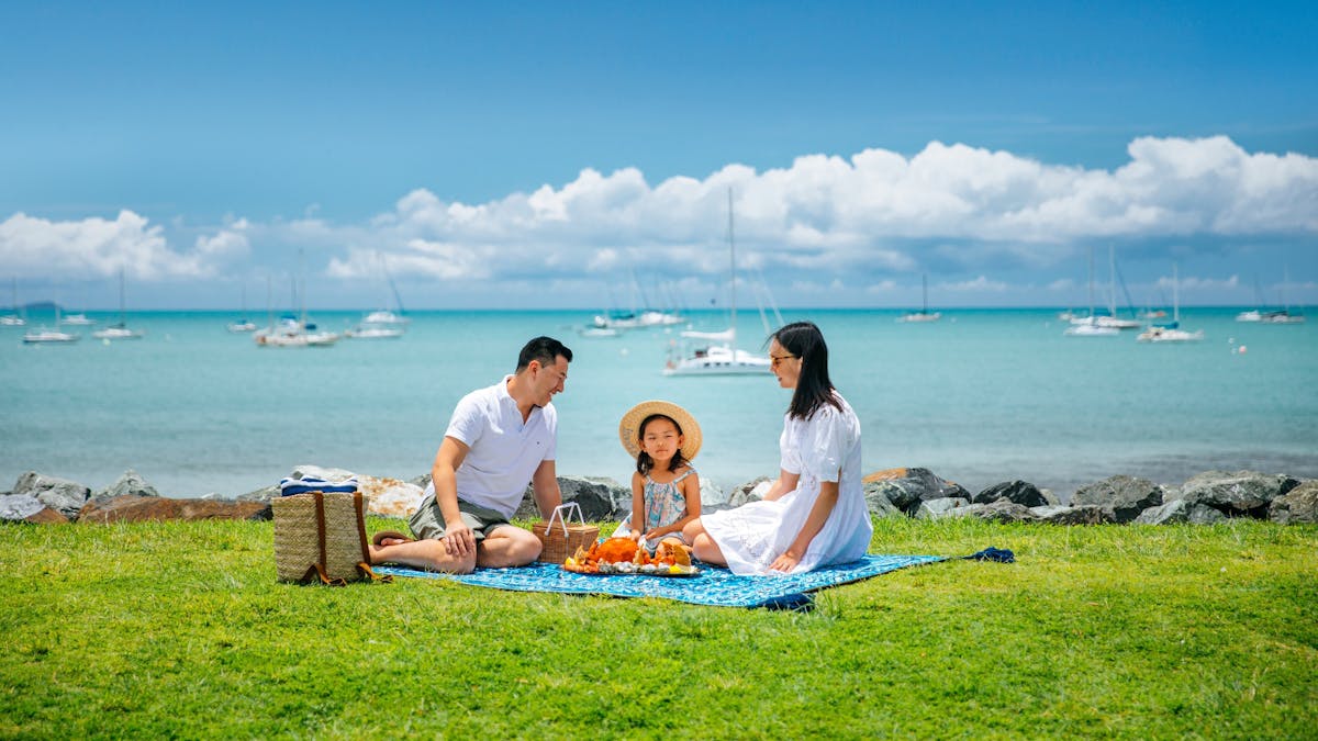 Couple and child sitting having a picnic on grass area with ocean and boats behind them