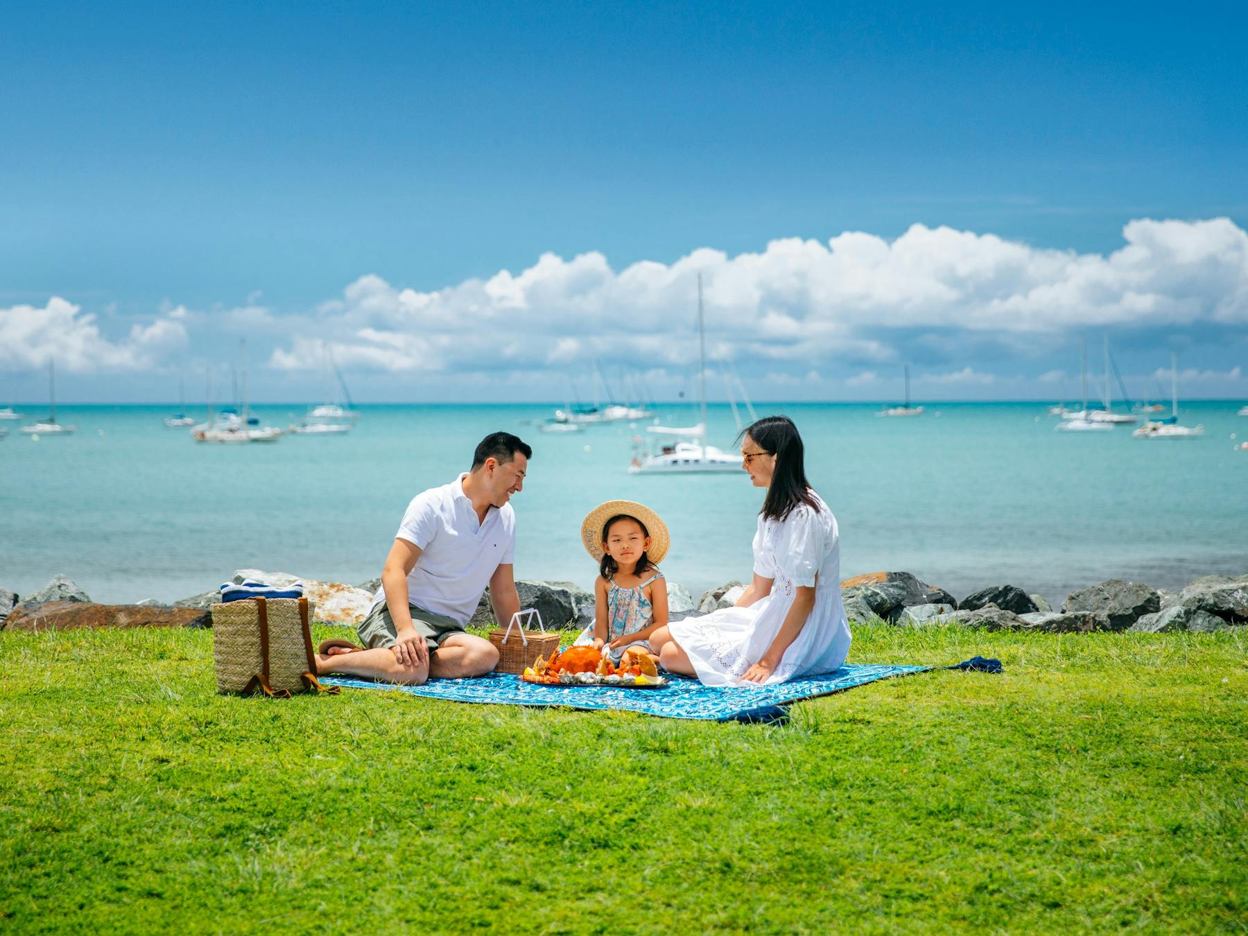 Couple and child sitting having a picnic on grass area with ocean and boats behind them