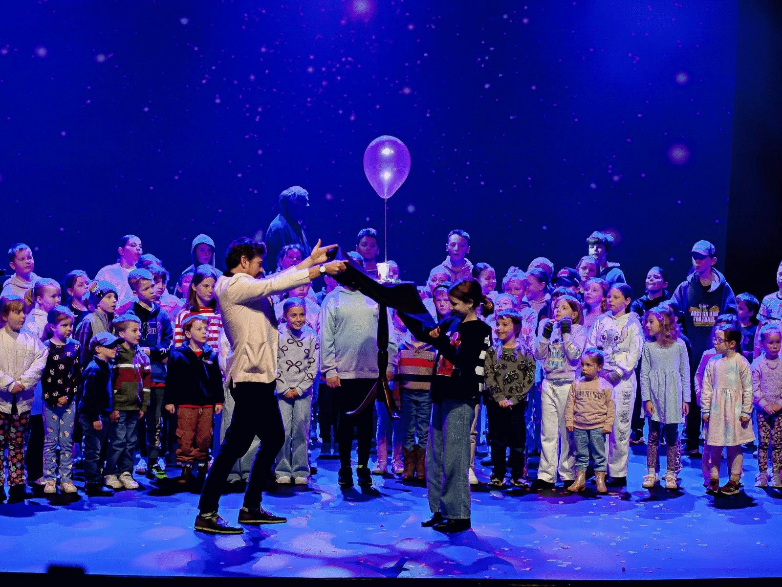 A large group of children and adults stand on stage, some watching two people in the center