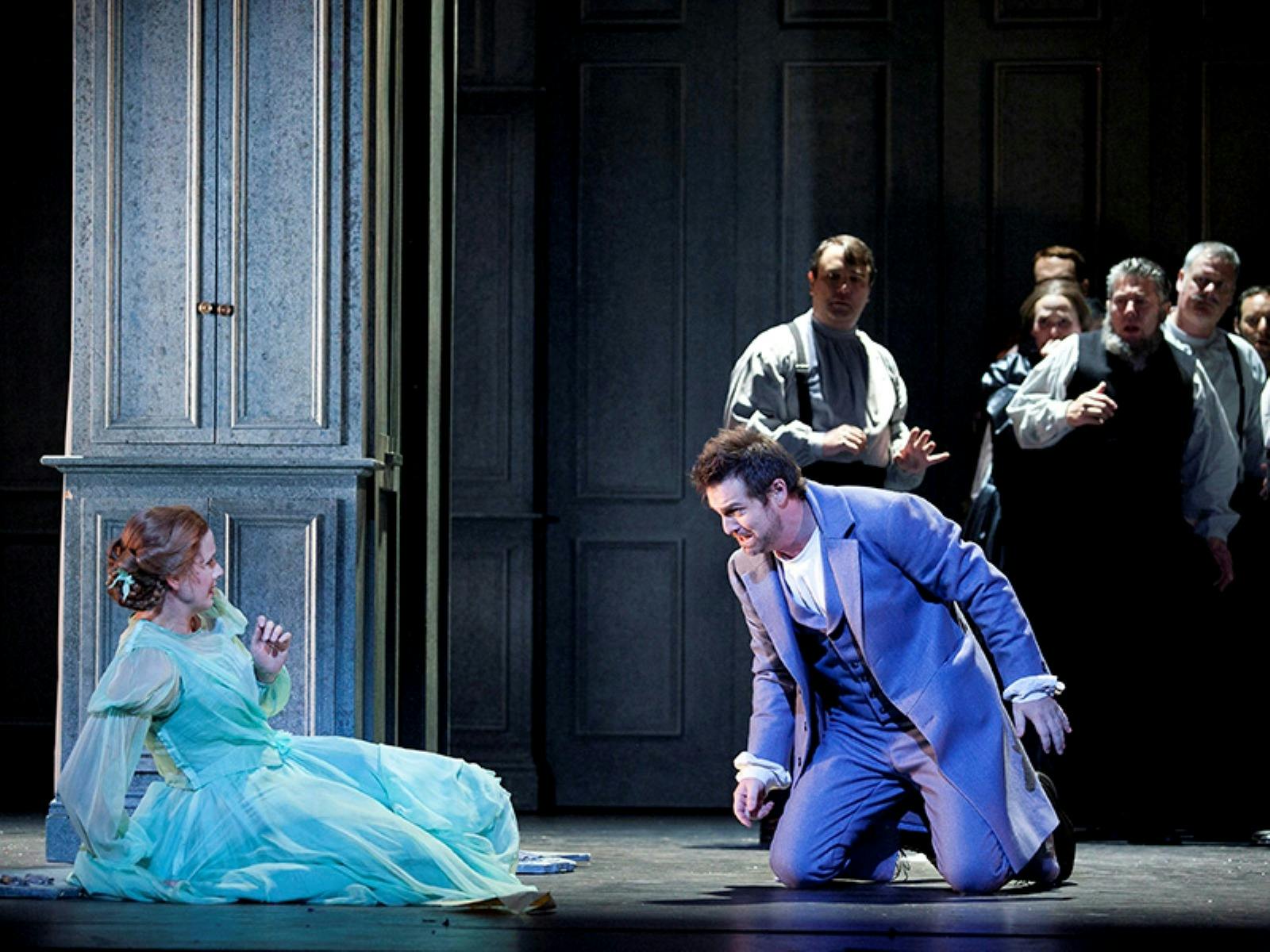 Kneeling man reaches toward woman in teal gown as onlookers watch in a dim stage setting