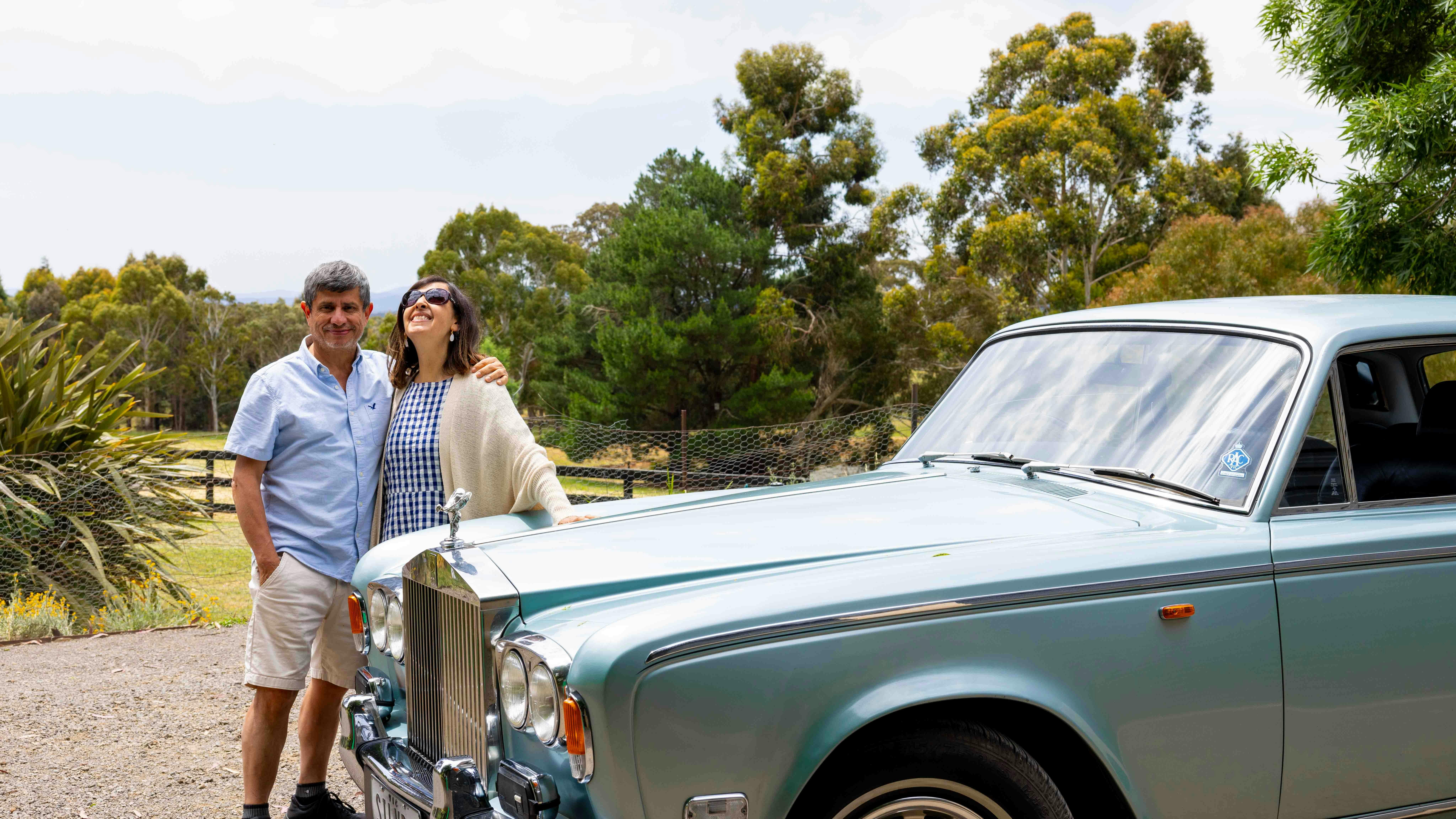 A couple from Colombia hug alongside the silver-blue Rolls Royce with trees behind them