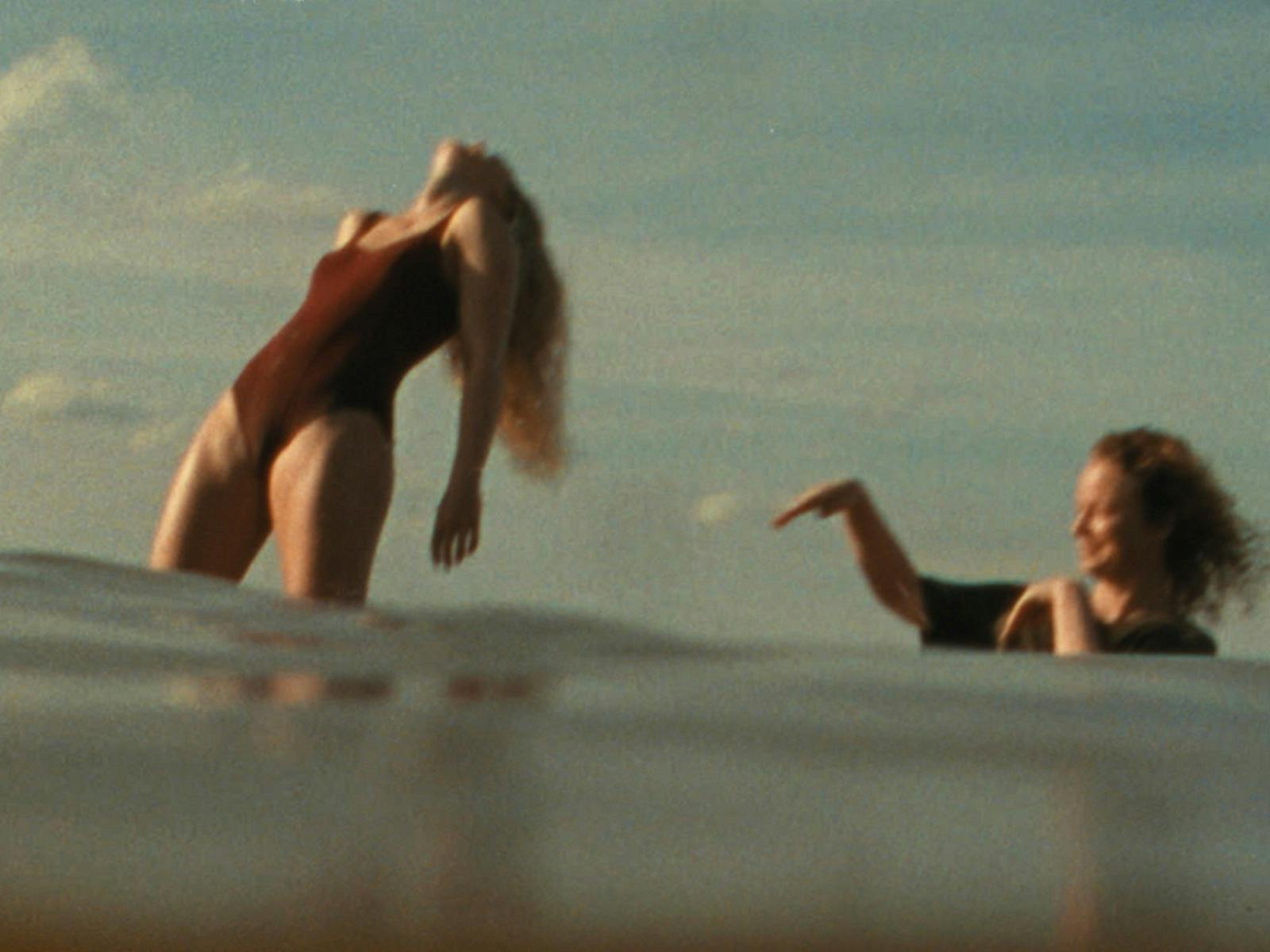 Woman in bathing suit standing in water leaning back with another woman flicking water at her
