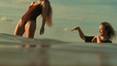 Woman in bathing suit standing in water leaning back with another woman flicking water at her